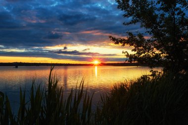 Gölün kıyısında gün batımı, ön planda ağaçlar ve sazlıklar.