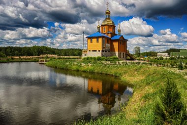Gölün kıyısındaki ahşap kilise, sudaki yansıma, mavi gökyüzü ve gri bulutlar.