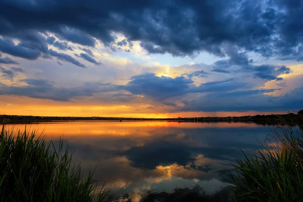 前景には雲と葦が描かれた穏やかな湖の上のカラフルな夕日の静かなシーン — ストック写真