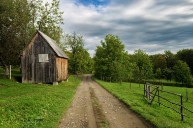 Dağ yamacındaki bir köy yolu ormana, ahşap bir eve, ahşap bir çite, mavi gökyüzüne ve beyaz bulutlara götürür.