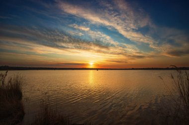 Nehir kıyısında güzel bir gün doğumu, renkli parlak bulutlar, ön planda sazlıklar.