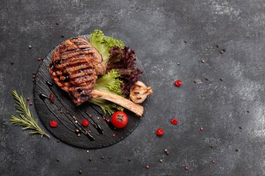 Kemiğin üstünde ızgara biftek, sarımsak ve siyah yuvarlak bir taşın üzerinde domates.