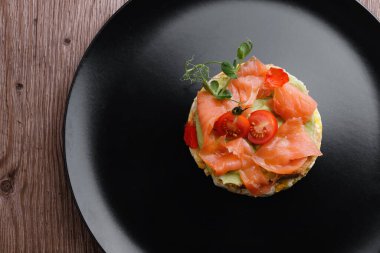 Kahvaltı, çırpılmış yumurtalı ekmek, somon, avokado ve domates, siyah bir tabakta.