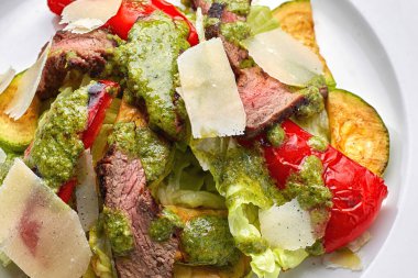 Dana eti, fırında sebze, dolmalık biber, kabak ve parmesan peynirli, beyaz tabakta, koyu bir arka planda.