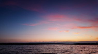 Sakin göl üzerinde dingin ve canlı bir günbatımı, sıcak renkleri gökyüzüne ve suya döküyor.