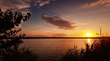 Sakin göl üzerinde huzurlu ve sıcak bir gün batımı, gökyüzünde altın renkler döküyor ve suyu yansıtıyor.