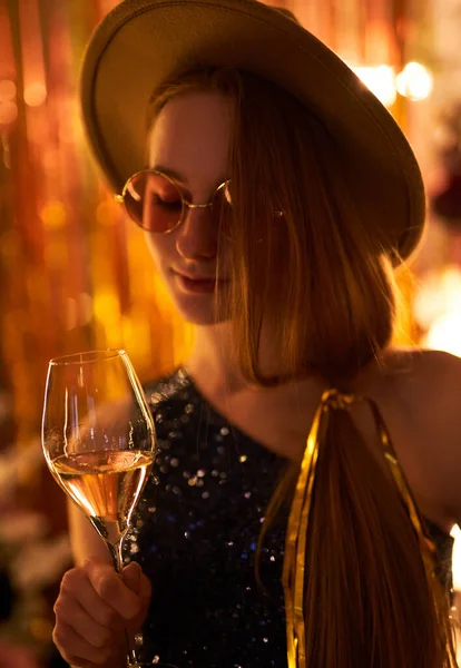 Happy and fun emotions during night party. Girl in hat and pink glasses holding glass of sparkling wine partying with blurred lights on background. High quality vertical image