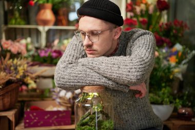 Yorgun küçük işletme sahibi konsepti. Çiçekçide kazak, bere ve gözlük takan beyaz bir çiçekçi. Kavanozda bitkileri olan yakışıklı bir sera sahibi ya da bahçıvan.