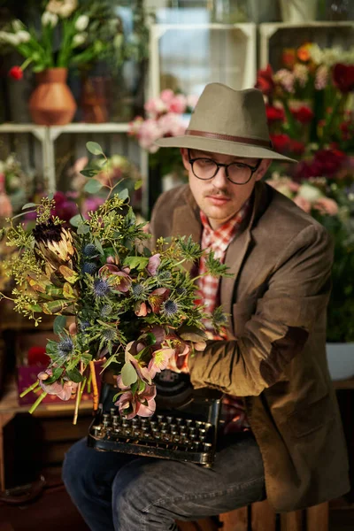 Tired or thinking emotion. Attractive caucasian male person in eyeglasses and hat sitting with old typewriter in flower shop. Poet or writer working concept. High quality vertical image