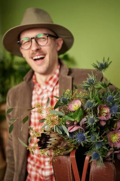 Attractive laughing male person in eyeglasses, vintage clothes and hat staying with flower bouquet and briefcase in flower store. Looking at camera. High quality vertical image