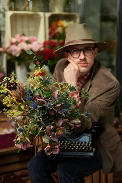 Poet or writer thinking concept. Cheerful caucasian male portrait in eyeglasses and hat sitting with old typewriter in flower store. High quality photo