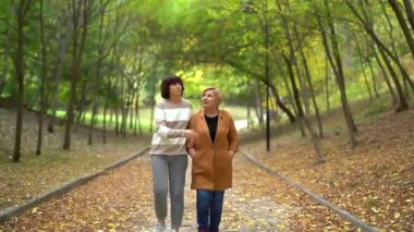 Anneler Günü konsepti. Kıdemli emekli anne ve yetişkin neşeli kızının portresi. Parkta, sonbahar sezonunda. Anne ve kızı sonbahar parkında yürüyor. Yüksek kalite 4k video