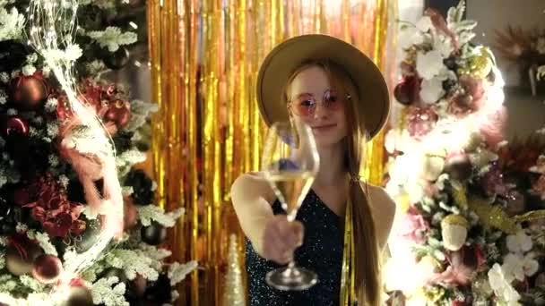 圣诞晚会上的快乐情绪 除夕之夜 戴着帽子和粉色眼镜的女孩端出一杯闪闪发光的葡萄酒或香槟 背景装饰着圣诞树 4K镜头 — 图库视频影像