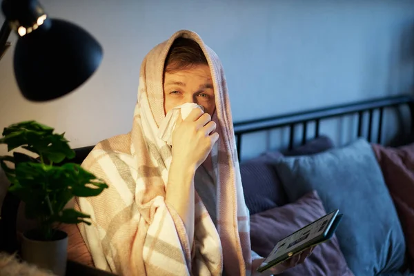 Feeling sick or illness concept. Distant work from home office or online education. Bearded young caucasian male using tablet in bed full covered with blanket. High quality image