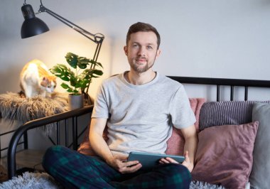 Çekici sakallı, serbest çalışan erkek yazılım mühendisi evde oturup tablet kullanıyor ve yakınlarda tekir kedisi oturuyor. Gri tişörtlü genç, yakışıklı müteahhit. Yüksek kaliteli resim