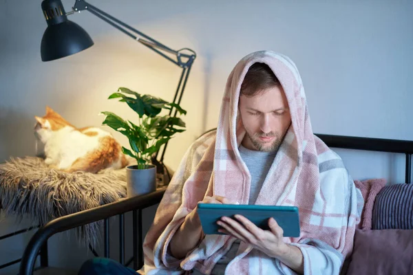 Caucasian man freelancer full covered with blanket using digital tablet in bed and white tabby cat sitting near him. Distant work from home office or online education. Feeling sick or illness concept