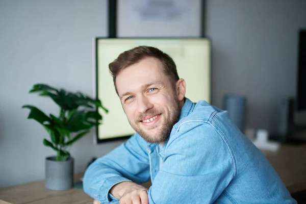 背景に肯定的な感情とコンピュータを持つデニムシャツのかわいい魅力的な白人男性 カメラを見て自宅のオフィスに座っている陽気な髭の男のフリーランサー笑顔 高画質 — ストック写真