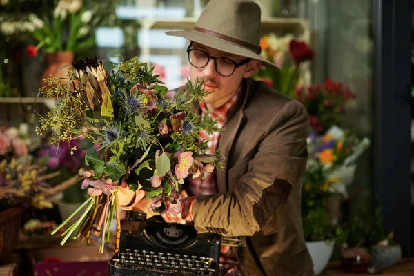 Cute retro styled caucasian male person in eyeglasses and hat sitting with old typewriter in flower shop. Poet or writer working concept. High quality photo