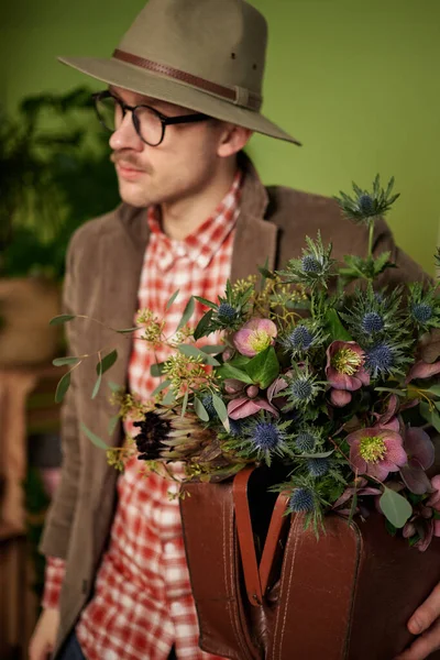 Attractive vintage styled male person in eyeglasses, velvet jacket and brown hat staying with flower bouquet and briefcase in flower store. High quality vertical image