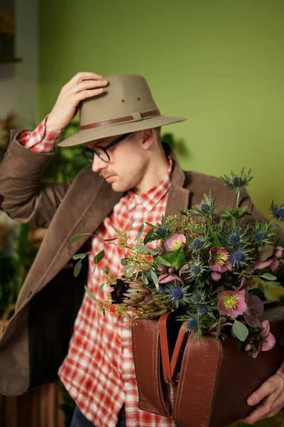 Attractive vintage styled male person in eyeglasses, velvet jacket and briefcase staying with flower bouquet correcting his hat in flower store. Poet or writer person concept. Vvertical image