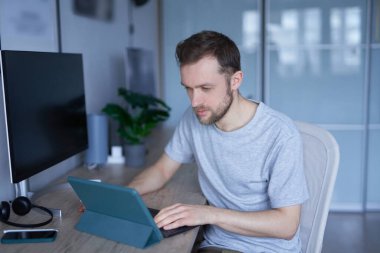 Çekici sakallı erkek evde oturup tablet kullanıyor ya da internetten eğitim veriyor. Gri gömlekli genç yakışıklının portresi. Yüksek kalite fotoğraf