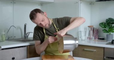 Çekici sakallı, yeşil önlüklü erkek aşçı plastik spatula kullanarak metal kaseden tahta kesme tahtasıyla hamur çıkarıyor. Ev mutfağı konseptinde çalışmak. Yüksek kalite 4k video kaydı