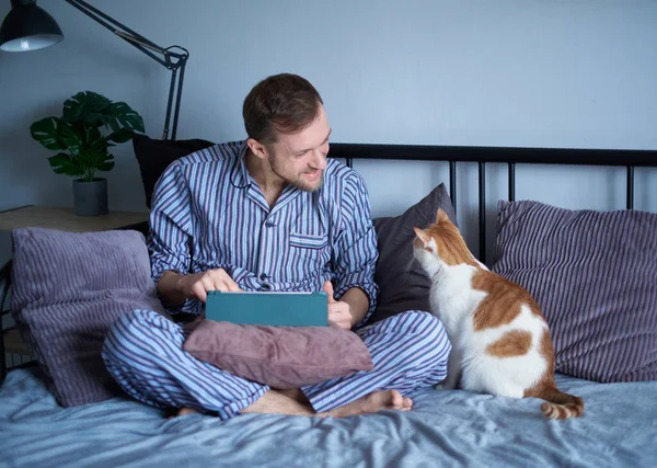 침대에 누워서 타블렛 컴퓨터를 사용하는 매력적 턱수염의 남자는 이그와 온라인으로 — 스톡 사진