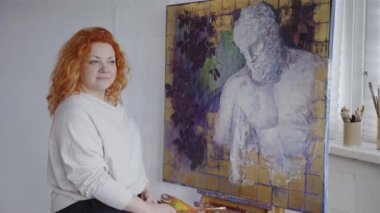 İlham konsepti. Kameraya bakan beyaz saçlı, çekici bir kadın sanatçının portresi. Sanat eserlerinin önünde poz veren kadın ressam. Yüksek kalite 4k video kaydı