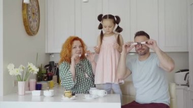 Aile hafta sonu konsepti. Neşeli beyaz anne babalar ve tatlı küçük kız sabahları kahvaltıda eğleniyor, ev mutfağındaki yemek masasında birlikte kurabiye yiyorlar. 4k video kaydı