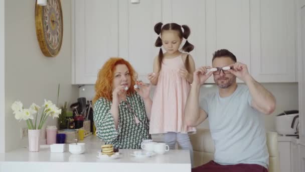 家族の週末のコンセプト 明るい白人の両親とかわいい小さな子供の女の子は 午前中に楽しい朝食をとり 家庭のキッチンのダイニングテーブルで一緒にクッキーを見ています 4Kビデオ映像 — ストック動画