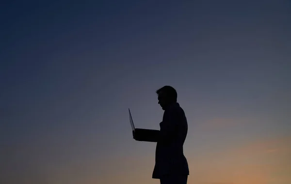 Male developer freelancer or it engineer working outdoor using laptop in silhouette at sunset. Hardly working evening. Adorable dusk at background. High quality image