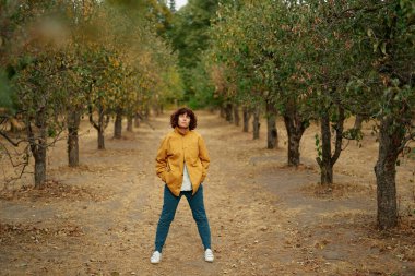 Sonbahar parkında sonbahar sezonunda neşeli kıvırcık kız. Turuncu ceketli, kot gömlekli ve pantolonlu, dik duran ve kameraya bakan kadın portresi, havanın keyfini çıkarıyor. Yüksek kaliteli resim