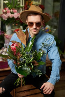 Sevgililer Günü ya da Uluslararası Kadınlar Günü konsepti. Güneş gözlüklü ve şapkalı mutlu hippi erkek çiçek dükkanında büyük taze bahar çiçeği buketi tutuyor. Yüksek kaliteli resim