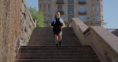 Adorable caucasian girl in jeans jacket running down on staircase holding flag of Ukraine. The concept of freedom or patriotism. Girl with the blue-yellow Ukrainian flag. 4k video footage