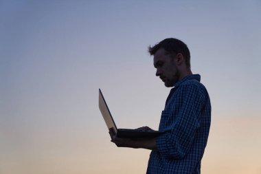Hardly working evening concept. Adorable developer freelancer or it male engineer standing working outdoor using laptop in silhouette at sunset. Beautiful dusk at background. High quality image