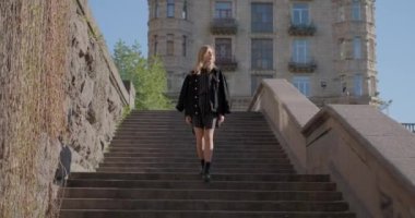 Adorable caucasian blonde girl in black jeans jacket going down on staircase on camera in European city center, springtime. High quality 4k video footage