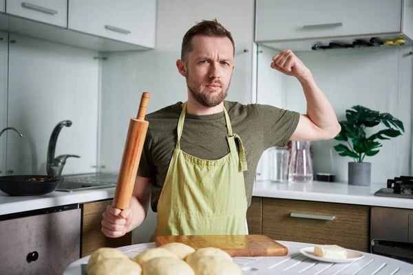 Handsome male baker at home sitting at the table with rolling pin. Portrait of serious faced caucasian baker man in green apron uniform looking at camera showing biceps muscles. High quality photo