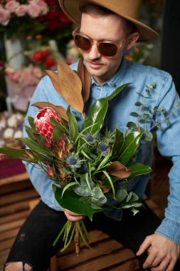 Valentines Day or International Womens Day concept. Adorable caucasian hipster male person in sunglasses and hat holding big fresh spring flower bouquet in flower store. High quality vertical image