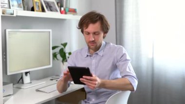 Closeup bearded caucasian male using tablet. Sitting on a chair with digital tablet relaxing and smiling in office with computer and bright background. Messaging, browsing the web, online shopping. 4K