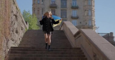 Adorable caucasian girl in jeans jacket running down on staircase holding flag of Ukraine. The concept of freedom or patriotism. Girl with the blue-yellow Ukrainian flag. 4k video footage