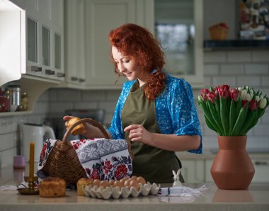 Woman smiling, preparing Easter basket with embroidered towel at kitchen home with tulips standing on the table. High quality photo