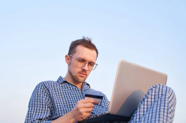 手にクレジットカードで買い物にノートを使用してかわいい白人男性 メガネと睡眠中の陽気な男は 午前中にノートパソコンの屋外を使用して着用オンラインで購入する — ストック写真