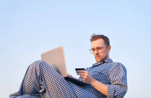 午前中のオンラインショッピング メガネの白人ひげを生やした男性は ノートパソコンやナイトウェアを着てクレジットカードを使って屋外で買い物をしています 背景に青空が広がっています 高画質 — ストック写真