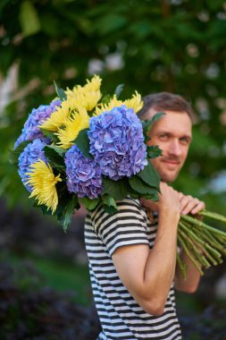 Elinde bir buket ortanca ve sarı çiçeklerle gülümseyen bir adam yeşil bahçenin arka planındaki kameraya bakıyor. Yüksek kalite fotoğraf