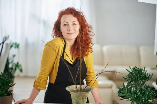 Female caucasian redhead florist portrait in yellow dress and black apron. Vintage vase with branches on foreground. Florist, plants care and gardening concept. High quality vertical image