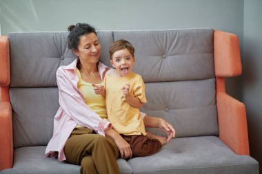 Evdeki kanepede oturan küçük çocukla oynayan beyaz bir kadının portresi. Aile zamanı kavramı. Yüksek kaliteli dikey fotoğraf