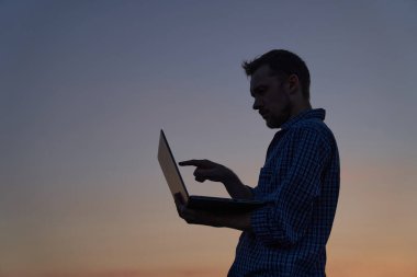 Çevrimiçi buluşma konsepti. Gün batımında ekranda parmaklarını siluet şeklinde gösteren dizüstü bilgisayar kullanarak dışarıda çalışan sevimli serbest çalışan erkek mühendis ya da bilişim destekçisi. Yüksek kaliteli resim