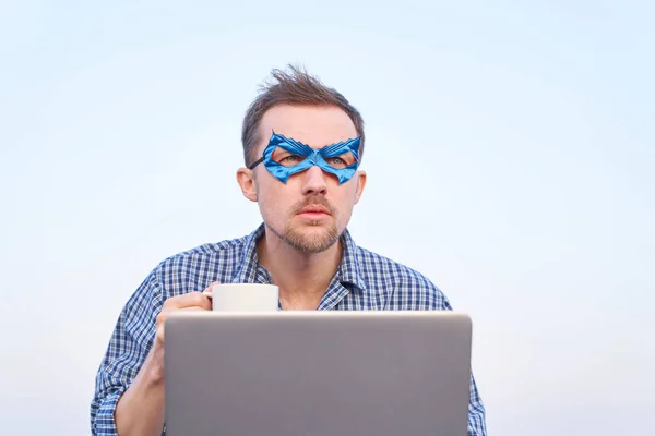 ナイトウェアのシャツを着た陽気な男とノートパソコンを屋外で使用して青い顔のマスク Itサポートスーパーヒーロー男ホットティーやコーヒーのノートパソコン保持カップを使用しています 高画質 — ストック写真