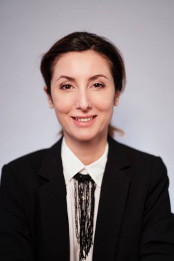 Siyah ceketli, gri arka planda poz veren gülümseyen esmer, beyaz iş kadını. Koyu takım elbiseli neşeli yönetici kadın gülüyor ve kameraya bakıyor. Yüksek kaliteli dikey fotoğraf