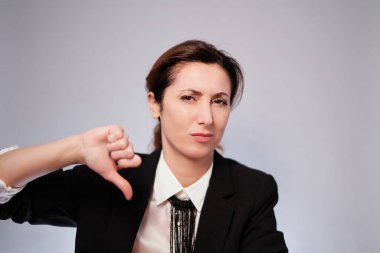Kadın negatif jest yapıyor. Siyah ceketli ciddi bir iş kadını gri arka planda poz veriyor. Koyu renk takım elbiseli kadın kameraya negatif yüzlü bakıyor. Yüksek kalite fotoğraf